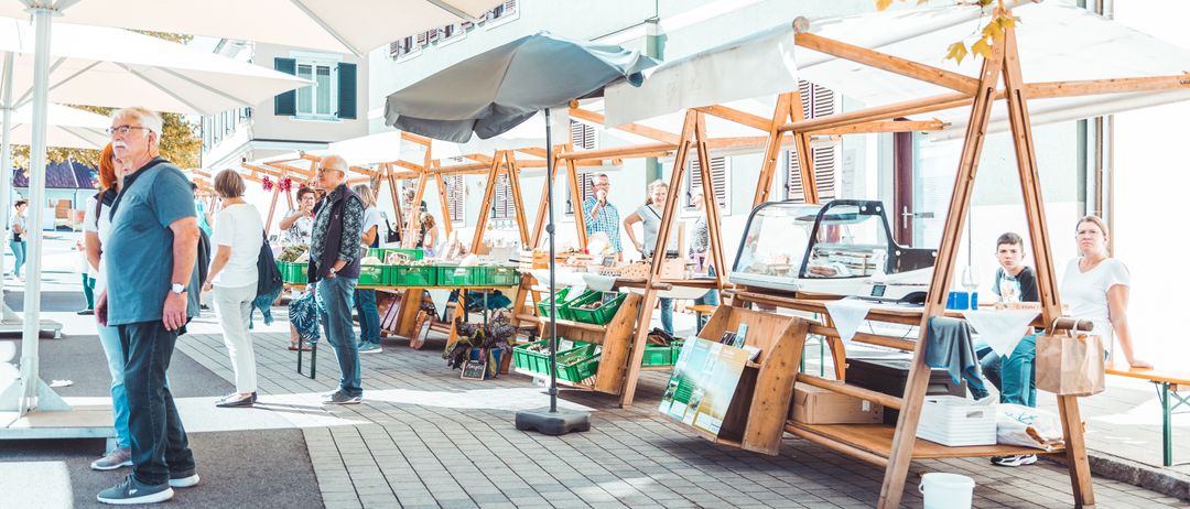 Ein Markt mit mehreren Ständen, darunter einer mit einer Frau hinter der Theke. Die Stände stehen unter großen Sonnenschirmen und Holzdächern, einige haben grüne Kisten. Leute gehen herum und interagieren mit den Händlern.