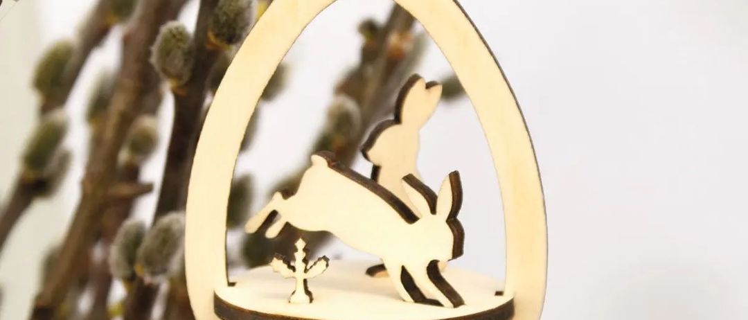 Eine hölzerne Osterdekoration in Form eines Eies mit einem springenden Hasen darin. Die Dekoration wird von einem Faden aufgehängt und ist von Zweigen umgeben.