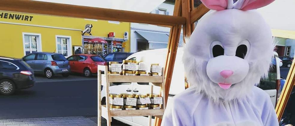 Eine Person in einem Hasenkostüm, die einen Korb mit Ostereiern hält, steht vor einem Tisch mit Lebensmitteln. Hinter ihr steht ein Schild mit der Aufschrift 'WALHALL Genusskulisse'. Im Hintergrund befindet sich ein gelbes Gebäude mit einem Geschäft und geparkten Autos.
