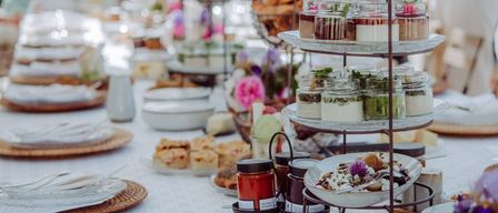 Ein langer Tisch, der für eine Party mit verschiedenen Desserts, Blumen und Utensilien gedeckt ist. Einige Menschen stehen hinter dem Tisch, einige tragen Sonnenbrillen.