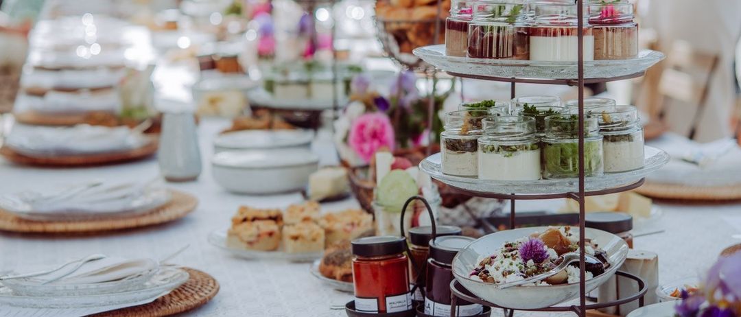 Ein langer Tisch, der für eine Party mit verschiedenen Desserts, Blumen und Utensilien gedeckt ist. Einige Menschen stehen hinter dem Tisch, einige tragen Sonnenbrillen.