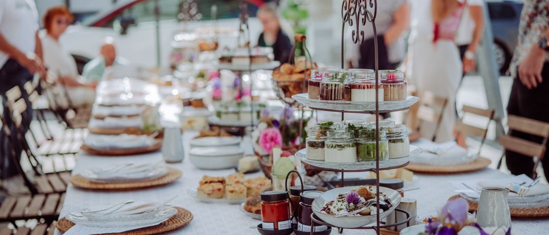 Ein langer Tisch ist mit Tellern, Schüsseln und dekorativen Gegenständen gedeckt. Es gibt kleine Teller mit Desserts und einen dreistufigen Ständer mit verschiedenen Desserts. Hinter dem Tisch stehen Menschen.
