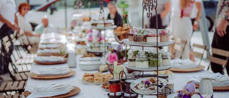 Ein langer Tisch ist mit Tellern, Schüsseln und dekorativen Gegenständen gedeckt. Es gibt kleine Teller mit Desserts und einen dreistufigen Ständer mit verschiedenen Desserts. Hinter dem Tisch stehen Menschen.