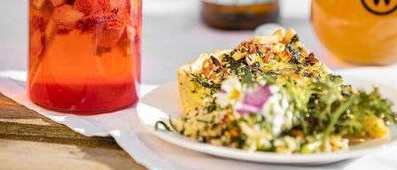 Ein Glas mit einem Erdbeergetränk, eine Flasche Bier und ein Glas Bier mit schaumiger Krone. Ein Teller mit einem Gemüsegericht und einer Torte steht daneben.