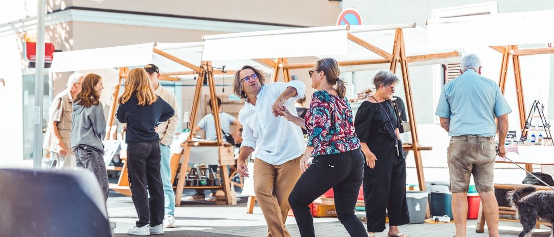 Ein Mann und eine Frau tanzen auf einem Markt, umgeben von Ständen und Menschen, während andere beobachten und mit der Umgebung interagieren.
