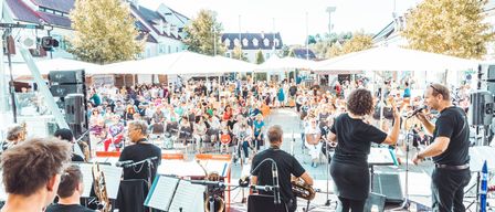 Eine Jazzband spielt auf einer Bühne vor einem großen Publikum unter Sonnenschirmen auf einem Stadtplatz.