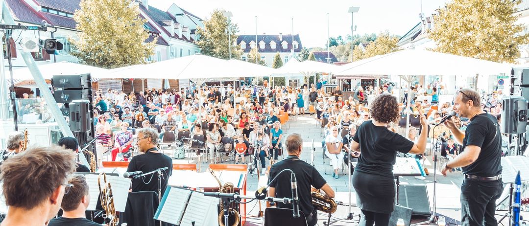 Eine Jazzband spielt auf einer Bühne vor einem großen Publikum unter Sonnenschirmen auf einem Stadtplatz.
