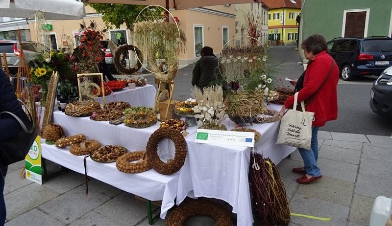 Eine Frau in einem roten Mantel durchstöbert einen Outdoor-Markt mit verschiedenen Lebensmitteln, Blumen und Kränzen unter einem Regenschirm. In der Nähe erkunden Menschen den Markt, und Autos sind entlang der Straße geparkt.