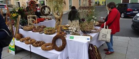 Eine Frau in einem roten Mantel durchstöbert einen Outdoor-Markt mit verschiedenen Lebensmitteln, Blumen und Kränzen unter einem Regenschirm. In der Nähe erkunden Menschen den Markt, und Autos sind entlang der Straße geparkt.