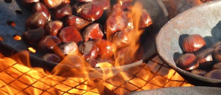 Drei Metallschüsseln voller gerösteter Kastanien stehen über einem offenen Feuer, das die Szene erhellt.