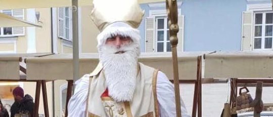 Ein Mann, der als Bischof verkleidet ist, steht auf einem Bürgersteig und hält einen Stab. Er trägt einen weißen Umhang mit goldener Stickerei und eine Mitra. Hinter ihm befinden sich Marktstände und geparkte Autos.