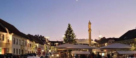 Ein belebter Stadtplatz mit geparkten Autos, einem Weihnachtsbaum und einem Halbmond am Himmel. Der Bereich ist von Straßenlaternen und Schildern beleuchtet.