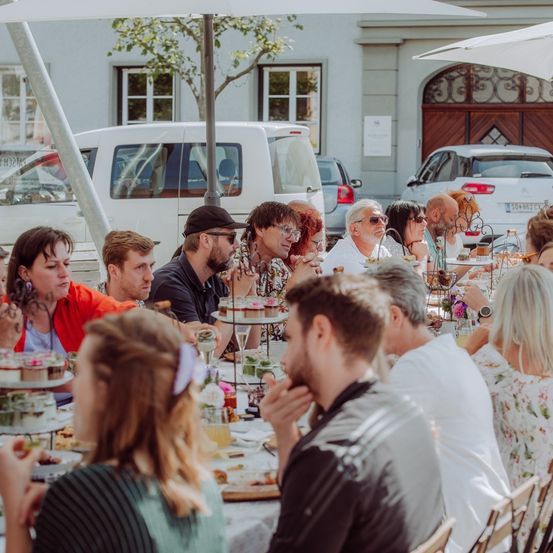 Eine Gruppe von Menschen sitzt an einem Tisch in einem Outdoor-Essbereich und genießt eine Mahlzeit. Der Tisch ist mit verschiedenen Gerichten beladen, und einige Leute essen, während andere in Gespräche vertieft sind. Im Hintergrund befinden sich Gebäude, Autos und Bäume.