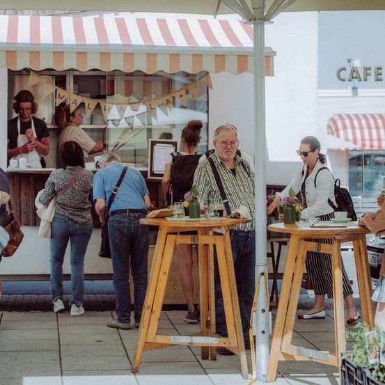 Ein Street-Food-Truck mit gestreiftem Vordach, Kunden an hölzernen Tischen und einem Schild für Cafe im Hintergrund.