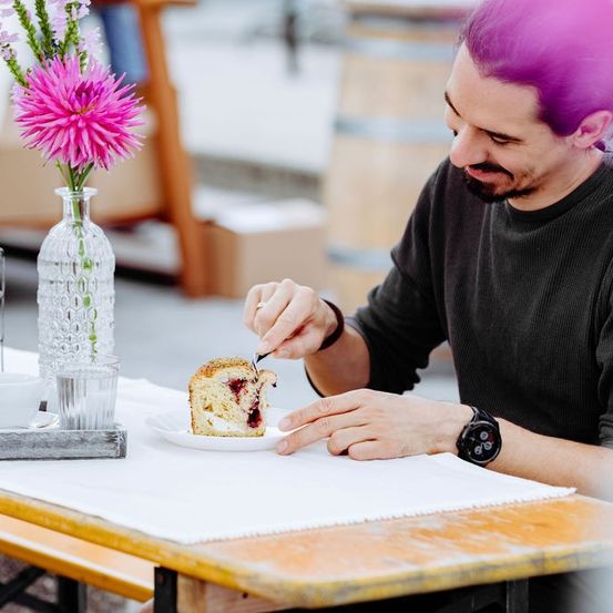 Ein Mann mit lila Haaren sitzt an einem Tisch und isst ein Stück Erdbeer-Sahne-Kuchen.