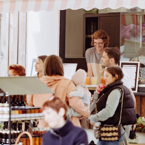 Ein Mann mit Brille lächelt und hält ein Baby, während er hinter einem Tresen steht. Hinter ihm hält eine Frau einen Becher und schaut zu ihm. Vor dem Tresen steht eine Gruppe von Menschen, darunter eine Frau, die ein Baby hält.