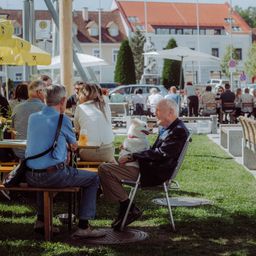 Ein Mann mit einem Hund auf dem Schoß sitzt auf einem Stuhl in einem Café im Freien, umgeben von anderen Leuten, die auf Bänken und Tischen sitzen.