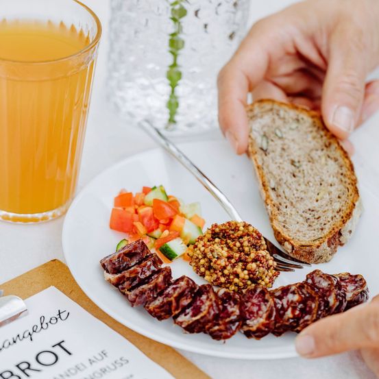 Nahaufnahme einer Person, die einen Teller mit Essen mit einer Gabel, einem Stück Brot und einem Glas Saft auf einem Tisch isst.