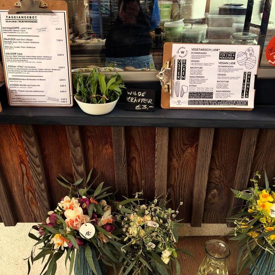 Eine hölzerne Theke mit einem Menübrett zeigt eine Auswahl an vegetarischen und veganen Gerichten. Das Menü umfasst verschiedene Salate, Suppen und Desserts, mit aufgeführten Preisen. Die Theke ist mit frischen Blumen und einer Topfpflanze geschmückt.