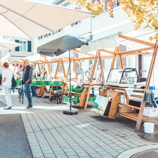 Menschen durchstöbern verschiedene Stände auf einem Markt. Ein Mann steht vor einem Stand, der verschiedene Artikel anbietet, während andere im Hintergrund sind.