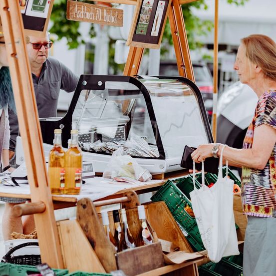 Ein Marktstand im Freien mit einem Schild, das 'Abbuchung 70 Biohof' liest. Ein Mann hinter dem Tresen schaut eine Frau an, die eine Tasche trägt. Verschiedene Flaschen und Produkte sind ausgestellt.