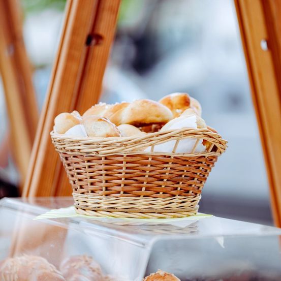 Ein Korb mit Brötchen ist auf einem transparenten Tisch ausgestellt, mit Holzrahmen darum. Die Brötchen sind ordentlich angeordnet, und einige sind mit weißem Tuch bedeckt.