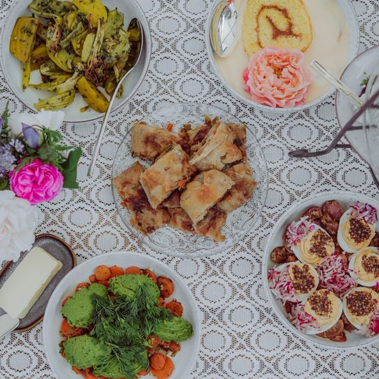 Ein Tisch mit verschiedenen Gerichten: gebratene Pasteten, Artischocken, gefüllte Eier und ein Dessert mit einem Zimtschnecken und einer rosa Rose.