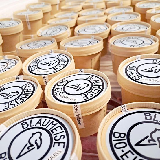 Eine Sammlung kleiner Behälter, jeweils mit einem Etikett, das den Markennamen 'BLAUMEISE', 'BIO-ESSEN' und 'WILDBEER' trägt, in einer Reihe auf einer Holzoberfläche angeordnet.