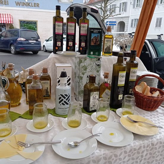 Ein Tisch zeigt verschiedene Flaschen Olivenöl, Gläser, Teller und einen Korb mit Brot. Mehrere Autos sind am Straßenrand geparkt.