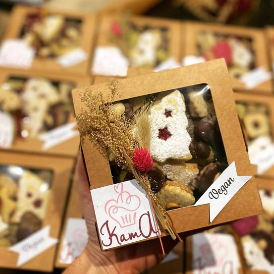 Eine Hand hält eine Schachtel mit verschiedenen Keksen und einer dekorativen Blumendekoration. Die Kekse sind vegan und werden in einem transparenten Fenster ausgestellt. Das Etikett lautet 'Kama Vegan.' Andere Schachteln sind im Hintergrund zu sehen.