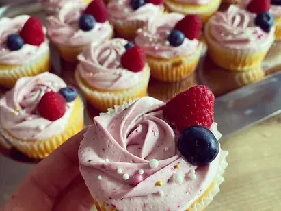 Eine Person hält einen Cupcake mit rosafarbener Glasur und einer Rosen-Design, gekrönt mit Himbeeren und Blaubeeren. Dahinter hält ein Tablett weitere Cupcakes, jeder mit ähnlicher Toppings und Glasur.