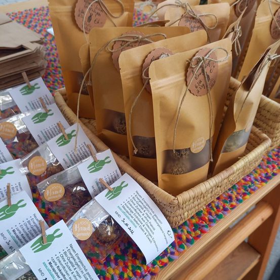Ein Korb voller brauner Papiertüten mit gebundenen Schnüren und Etiketten liegt auf einem bunten Tuch. Jede Tüte hat ein Etikett mit '100% Handgemacht' darauf. Die Tüten sind mit pflanzlichen Produkten gefüllt.