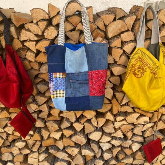 Drei Taschen hängen vor einem Hintergrund aus Feuerholz. Die mittlere Tasche ist ein Patchwork aus Denim und andere Taschen sind rot und gelb.