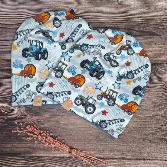 Zwei Mützen mit Traktormuster liegen auf einer Holzoberfläche. Jede Mütze hat Traktoren in verschiedenen Farben und Sternen.