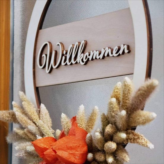 Ein dekorativer Holzkranz mit dem Schild 'Willkommen' ist an einer Wand befestigt und zeigt getrocknete Gräser und ein orangefarbenes Blatt in der Mitte.
