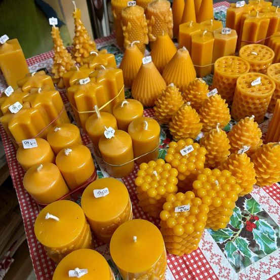 Ein Tisch ist mit einem rot-weiß karierten Tischtuch bedeckt. Er zeigt verschiedene gelbe Kerzen in unterschiedlichen Formen, wie Kegel, Tannenzapfen und Kugeln. Jede Kerze hat einen Preisanhänger. Der Hintergrund ist leicht verschwommen.