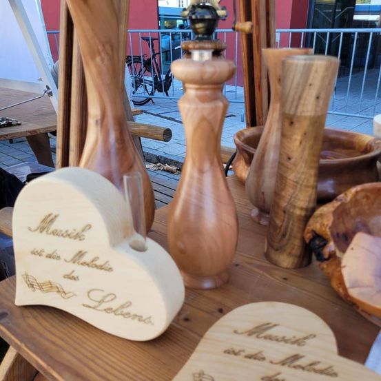 Auf einem Holztisch sind verschiedene geschnitzte Holzgegenstände ausgestellt, darunter ein herzförmiges Stück mit dem Text 'Musik ist die Melodie der Liebe.' Ein Pfeffermühle und andere Holzschüsseln sind ebenfalls vorhanden. Ein Fahrrad ist hinter einem Geländer geparkt.
