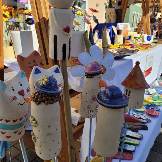 Eine Ausstellung von handgefertigten Keramik-Katzenfiguren und Blumenskulpturen auf einem Marktstand. Die Katzen tragen Hüte und die Blumen haben leuchtende Farben.