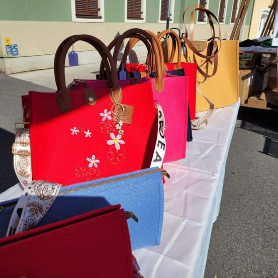 Eine Ausstellung von farbenfrohen Handtaschen mit Blumendesigns und Etiketten, auf einem weiß bezogenen Tisch vor einem Gebäude angeordnet.