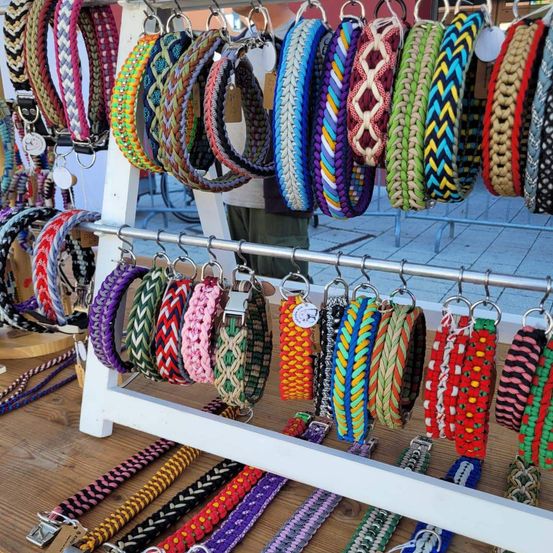 Ein Stand mit einer Vielzahl farbenfroher, geflochtener Armbänder und Halsbänder, die an Haken hängen und auf einem weißen Gestell über einer Holzoberfläche ausgestellt sind.