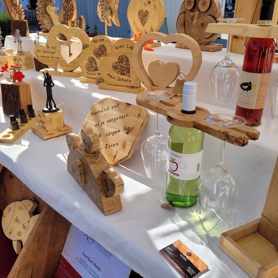 Eine Ausstellung von holzgeschnitzten herzförmigen Objekten, Weingläsern und Flaschen. Eine kleine Blume, Schlüssel und eine Visitenkarte sind ebenfalls vorhanden. Der Tisch ist mit einem weißen Tuch bedeckt.