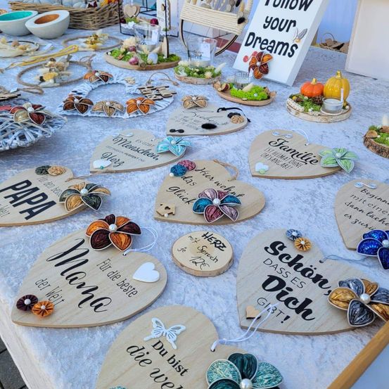 Auf einem Tisch sind verschiedene Holzherzen mit unterschiedlichen Botschaften zu sehen. Die Herzen tragen Aufschriften wie 'MAMA', 'VORSORGE' und 'SCHOEN'. Sie sind mit dekorativen Blumen und Schmetterlingen verziert. Im Hintergrund befinden sich weitere Dekorationsartikel wie Kürbisse und ein Schild mit der Aufschrift 'Follow Your Dreams'.