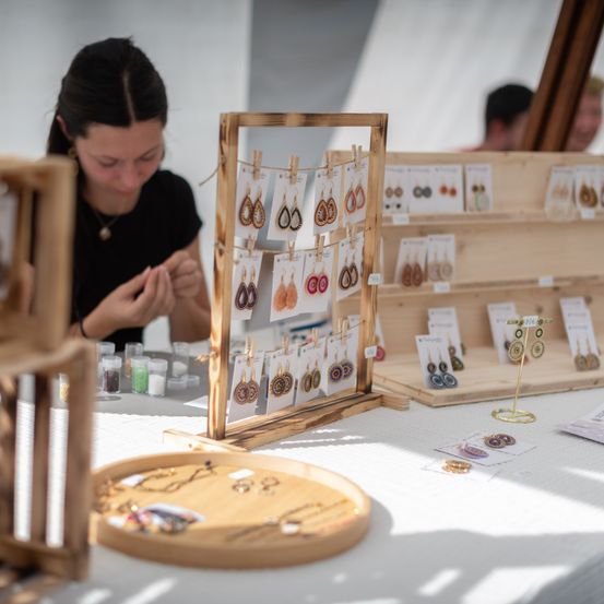 Eine Frau richtet einen Stand mit handgefertigten Ohrringen auf einem weißen Tisch ein, mit Holzkisten und Tabletts mit verschiedenen Accessoires.