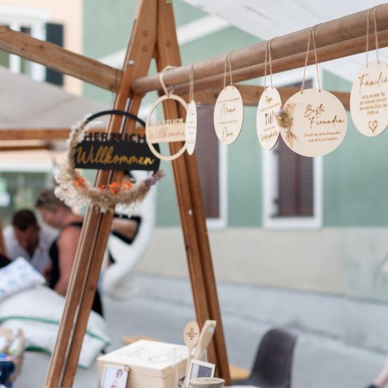 Eine Holzstruktur mit einem Kranz und einem 'Herzlich Willkommen'-Schild. Hängende Holzanhänger mit Namen und Blumen. Eine weiße Box mit einem Fotorahmen. Ein verschwommener Hintergrund von sitzenden und stehenden Personen.