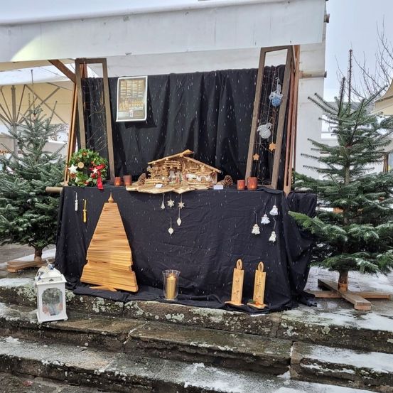 Eine Weihnachtsdekoration im Freien mit einer Krippe auf einem schwarz bespannten Tisch, umgeben von Kerzen, Ornamenten und Tannenbäumen.