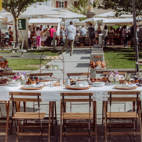Ein geschäftiger Outdoor-Markt mit mehreren Essensständen unter weißen Sonnenschirmen. Ein langer Tisch mit Essen und Getränken ist auf dem Bürgersteig aufgebaut. Leute gehen und shoppen.