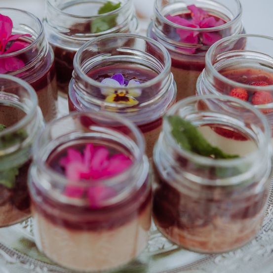 Mehrere klare Glasgläser mit geschichteten Desserts, jedes mit Blumen und Kräutern verziert, auf einem silbernen Tablett angeordnet.