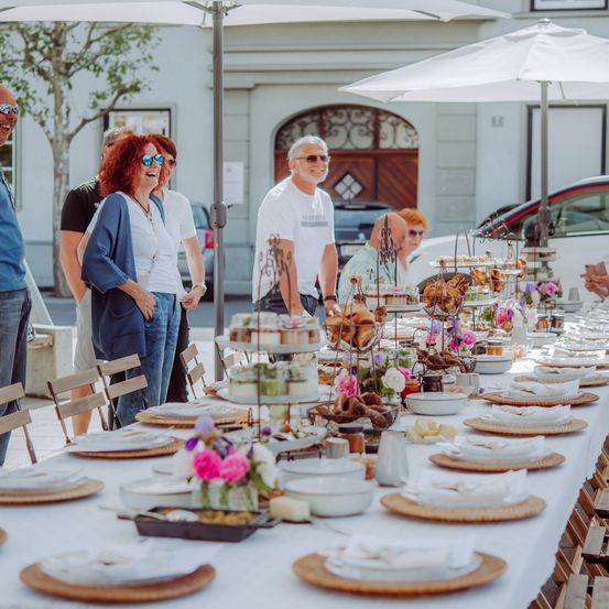 Ein langer Tisch für ein Treffen im Freien, mit Tellern, Blumen und verschiedenen Speisen unter Schirmen. Menschen in lässiger Kleidung stehen um den Tisch herum.