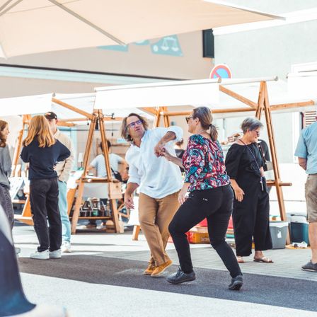 Mehrere Menschen tanzen im Freien unter einem Regenschirm. Hinter ihnen befinden sich Zelte und Stände, an denen einige Leute stöbern. Der Bereich scheint ein Markt oder Jahrmarkt zu sein.