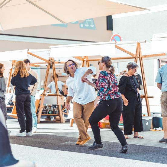Mehrere Menschen tanzen im Freien unter einem Regenschirm. Hinter ihnen befinden sich Zelte und Stände, an denen einige Leute stöbern. Der Bereich scheint ein Markt oder Jahrmarkt zu sein.